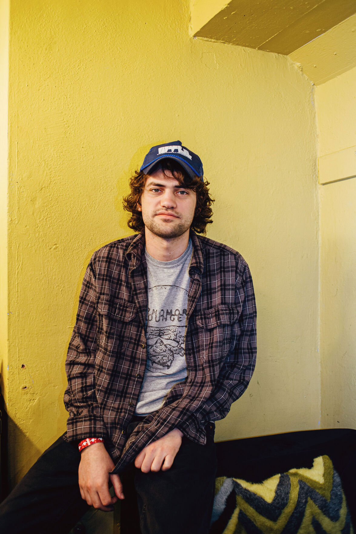 MJ Lenderman press photo artist sits on the arm of a striped couch in a room with yellow walls